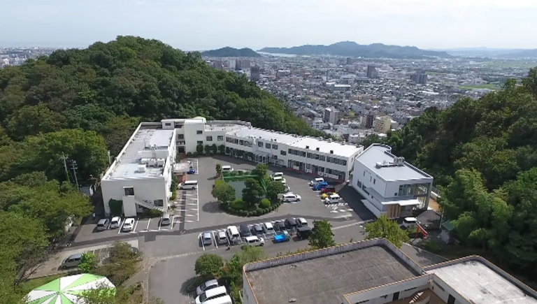 高台からの施設全体俯瞰。白い多棟建物と駐車場、周辺の山々と市街地が見える